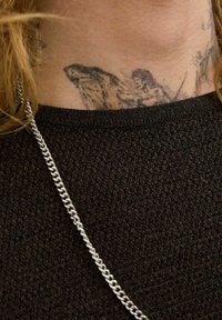 Fekete textúrájú pulóver gallér, ezüst láncnyaklánccal. A nyakon látható tetoválások részletes képeket ábrázolnak.