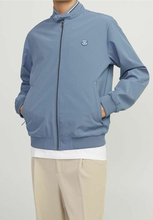 Homme portant une veste bomber bleue zippée avec des poignets côtelés et un pantalon beige, les mains dans les poches, debout devant un fond uni.