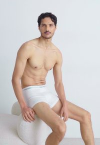 Boxers blancs en coton avec une ceinture élastique douce portant la marque "Calvin Klein", portés par un modèle masculin assis sur un tabouret blanc texturé.