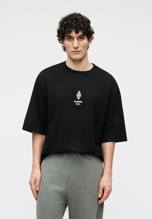 Noor noormehe lühikeste lokkis juustega seisab otse, kandes musta, üle suuruse T-särki, millel on keskel valge logo, ning hallid püksid.