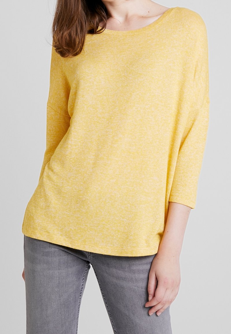 Femme portant un haut jaune ample à manches trois-quarts associé à un jean gris, debout devant un fond uni gris clair.