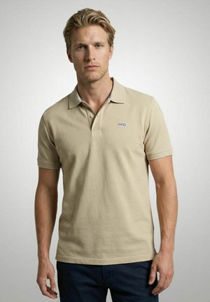 Blonde man die een beige korte mouwen polo met MCG-logo en een donkere broek draagt, staand tegen een effen lichte achtergrond.