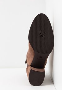 Bottine en daim marron avec une semelle en caoutchouc noir texturé, présentant un léger talon et un simple détail de boucle sur le côté.