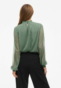 Grøn gennemsigtig bluse med lange, puffede ærmer, flæsekrave og en nøglehuldetalje bagpå, kombineret med sorte bukser. Glat tekstur.