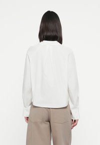 Weiße Bluse mit geradem Schnitt, langen Ärmeln und Knopfmanschetten. Verfügt über eine Rückenfalte und einen klassischen Kragen, kombiniert mit beigen Hosen.