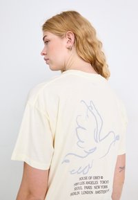 Geel katoenen t-shirt met een lichtgrijze duifafbeelding op de achterkant en tekst met de namen van verschillende steden gedrukt eronder. Korte mouwen, relaxte pasvorm.