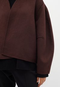 Ruda vilnonė apykaklė su laisvu siluetu, giliu V formos iškirpimu ir ilgomis rankovėmis; uždėta ant juodos raukšlėtos megztinio ir juodų kelnių.