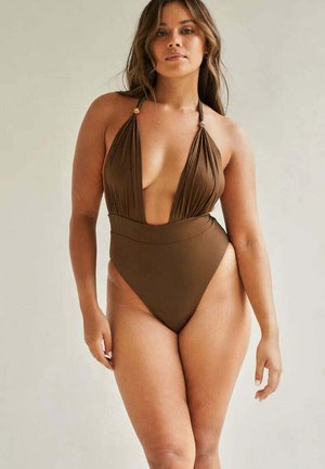 Kobieta ubrana w brązowy jednoczęściowy strój kąpielowy z głębokim dekoltem w serek i wiązaniem na szyi, stojąca na jednolitym jasnym tle.