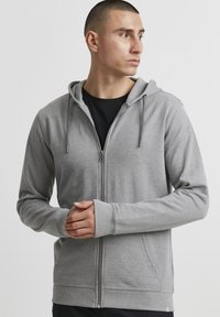 Sweat à capuche gris zippé en tissu doux, doté d'une capuche, de poches avant et de poignets côtelés. Design simple sans motifs ni logos visibles.