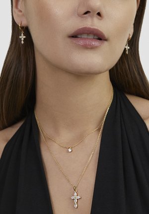 Close-up of a woman wearing gold cross earrings and layered gold necklaces with diamond-like pendants against a black halter top.