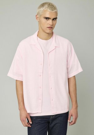 Chemise rose clair à manches courtes avec un devant boutonné, fabriquée en tissu texturé. Portée par-dessus un t-shirt rose clair assorti.