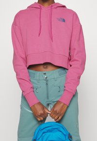 Sudadera rosa recortada con cordones y logo en el pecho, combinada con pantalones en tono verde azulado. Tela texturizada y detalles de puños acanalados visibles.