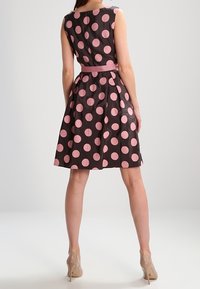 Vestido negro sin mangas con grandes lunares rosas, cintura entallada y falda acampanada. Combinado con un cinturón rosa y tacones neutros.