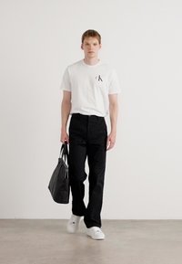 White t-shirt with a small black logo, black trousers, white sneakers, and a black tote bag. Smooth, casual fabric with a simple design.
