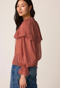 Blouse transparente rose avec des pois texturés, encolure froncée et poignets élastiqués, associée à un jean en denim bleu clair.