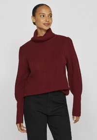Pull en maille bordeaux avec un col haut et replié, des côtes texturées et des manches légèrement bouffantes, associé à un pantalon noir.