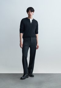 Un tânăr stă sprijinit de un perete simplu, purtând o cămașă neagră cu mâneci lungi, pantaloni negri și pantofi negri cu șireturi, privind înainte.