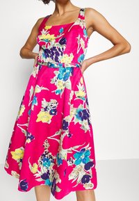Blommig klänning i stark rosa med blå, gula och lila mönster. Designad med fyrkantig halsringning och  åtsittande midja. Tillverkad av lättviktsmaterial.