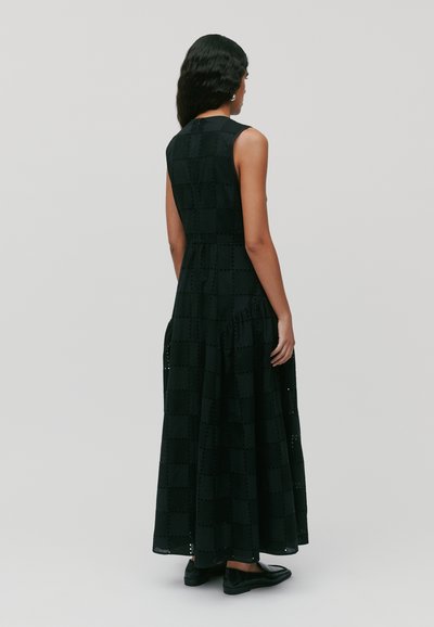 Schwarzes Maxikleid mit einem karrierten Muster, ausgestattet mit einem taillierten Oberteil, einem ausgestellteten Rock und Ausschnitten. Kombiniert mit schwarzen flachen Schuhen.