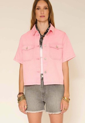 Femme portant une veste boutonnée à manches courtes rose clair sur un haut blanc, un short en jean gris, un foulard noir autour du cou et des bracelets dorés.