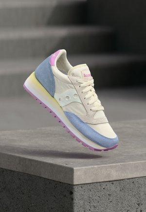 Kremasti tenisice s plavim i ružičastim akcentima te potplatom s žutim prugama, viseći iznad sive betonske ograde na otvorenom prostoru.