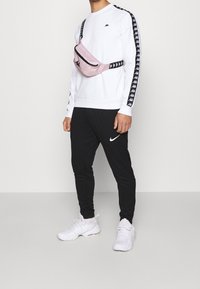 Witte sweatshirt met zwarte armstrepen, zwarte sportbroek en een roze heuptas die over het lichaam wordt gedragen. Witte sneakers maken de look compleet.