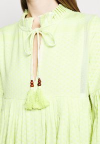 Světlé zelené šaty s texturovanou tkaninou, nařaseným výstřihem a klíčkovým předním dílem. Obsahují zavazování s třásněmi a akcenty ze dřevěných korálků.