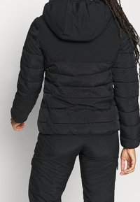 Svart puffjacka med huva, med en kviltad design som har horisontella sektioner, tillverkad av ett mjukt tyg, vy från baksidan.