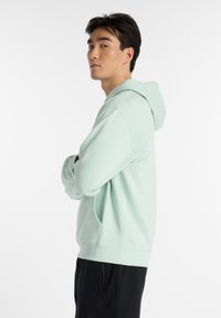 Sudadera verde menta con capucha ajustable, bolsillo tipo canguro y puños de canalé. Combinada con pantalones deportivos negros, que exhiben un ajuste holgado.