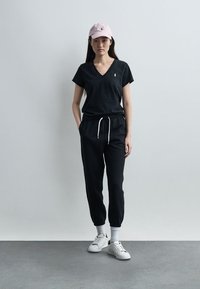 Γυναίκα με μαύρο μπλουζάκι V-neck και φόρμες, λευκά αθλητικά παπούτσια, λευκές κάλτσες και ροζ απαλό καπέλο baseball, στέκεται μπροστά σε απλό φωτεινό τοίχο.