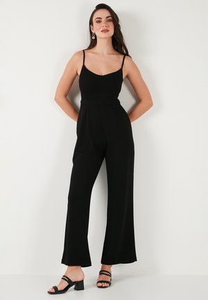 Mono negro sin mangas con escote en V, cintura ceñida y pantalones de pierna ancha. Tela suave, diseño sencillo, combinado con tacones negros de tiras.