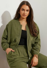 Giacca bomber verde oliva con chiusura a zip e due tasche frontali, abbinata a un top nero e pantaloni verde oliva coordinati, caratterizzati da una vestibilità rilassata.