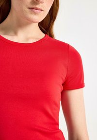 Jongere met rood haar en sproeten, gekleed in een aansluitend kort mouw rood T-shirt tegen een lichte achtergrond.