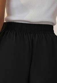 Black, pleated waistband of pants, featuring elastic material, worn with a white ribbed tank top that has a snug fit.