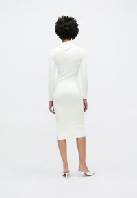 Λευκό πλεκτό φόρεμα με ριμπ υφή, μακριά μανίκια και γιακά. Σχεδιασμός μεσαίου μήκους με εφαρμοστή σιλουέτα, συνδυασμένο με μουλάρια.