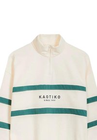 Sudadera de color crema con cuello alto, que presenta rayas en tono teal y el texto "KAOTIKO" con "DESDE 1999" debajo.
