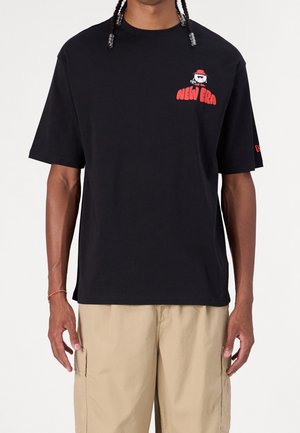 T-shirt noir New Era avec petit logo rouge représentant un chien portant des lunettes de soleil et un chapeau rouge, porté avec un pantalon cargo beige clair.