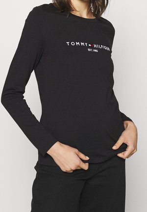 Kobieta ubrana w czarną bluzkę z długim rękawem z logo "Tommy Hilfiger EST. 1985", trzymająca rąbek, na jednolitym jasnym tle.