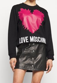 Schwarzer Sweatshirt mit einem leuchtend pinken Herzelement und dem Text "LOVE MOSCHINO" in Weiß. Kombiniert mit einem schwarzen Lederrock mit Druckknöpfen.