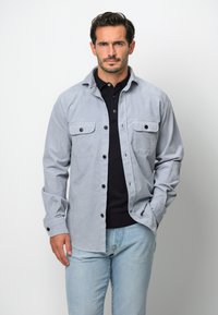 Ljusgrå skjorta med knappar, två bröstfickor och svarta knappar, lager på en svart polo, ihop med ljusblå jeans.