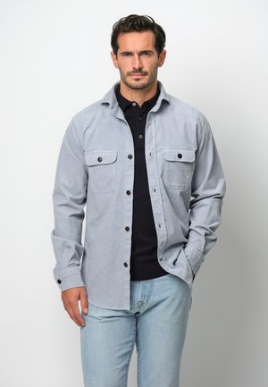 Hellgrau kariertes Hemd mit zwei Brusttaschen und schwarzen Knöpfen, über einem schwarzen Poloshirt getragen, kombiniert mit hellblauen Jeans.