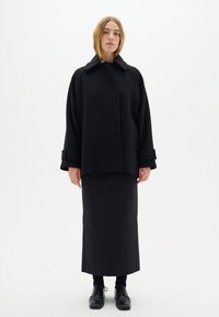 Zwarte wollen jas met een hoge kraag, voorzien van oversized mouwen en zakken, gecombineerd met een zwarte rechte rok en schoenen. Eenvoudige achtergrond.