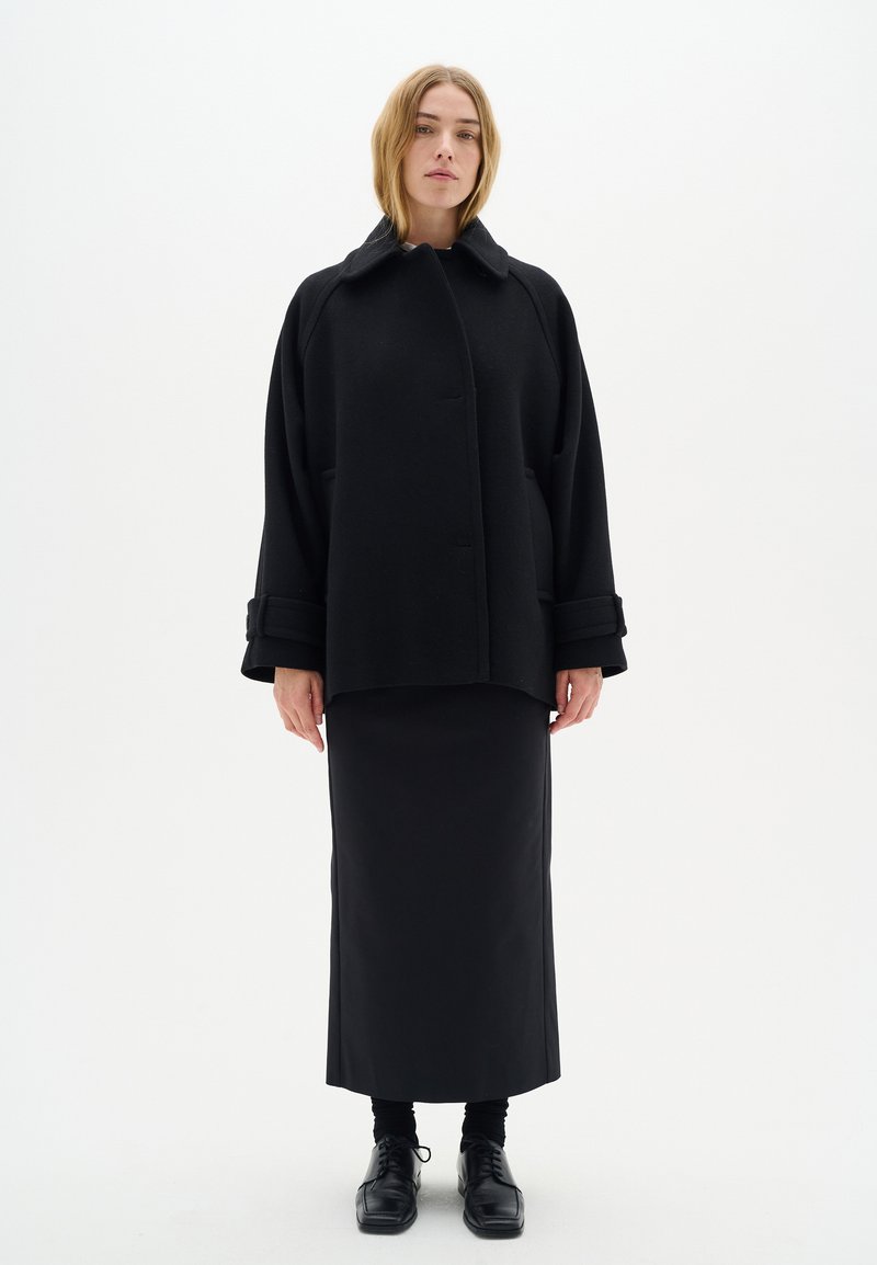 Zwarte wollen jas met een hoge kraag, voorzien van oversized mouwen en zakken, gecombineerd met een zwarte rechte rok en schoenen. Eenvoudige achtergrond.