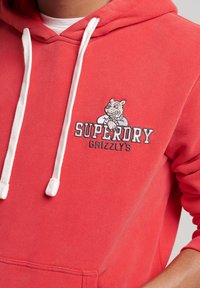 Felpa rossa con cappuccio realizzata in cotone con lacci bianchi. Presenta una stampa grafica di un orso e il testo "SUPERDRY GRIZZLY'S" in colori contrastanti.