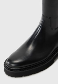 Bota de cuero negro con un acabado elegante, punta redondeada y suela gruesa de goma negra con un borde texturizado para un mejor agarre.
