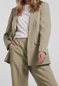 Femme portant une veste et un pantalon de costume beige avec un t-shirt blanc, la main dans la poche, sur un fond gris uni.