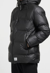 Schwarze Pufferjacke mit glänzender Oberfläche, dicker Polsterung, verstellbare Kapuze, Reißverschlusstaschen und einem Logotag am Saum.