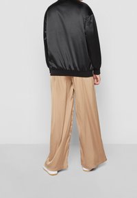 Veste bomber en satin noir avec une bande côte, associée à un pantalon large beige, montrant une texture lisse et une coupe décontractée. Des baskets blanches complètent le look.