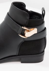 Černé kotníkové boty vyrobené z hladké kůže s lesklým zlatým sponou a plochým podpatkem. Mají zaokrouhlenou špičku a nízký střih.