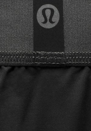 Svart atletisk tyg med en texturerad yta, ett kontrasterande slätt band och en rund logotyp. Sömmarna visar detaljerad sömnad.